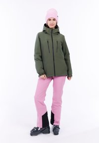 Grüne wasserdichte Jacke mit schwarzen Reißverschlüssen; rosa Hose mit schwarzen Akzenten; rosa gestrickte Mütze; schwarze Skistiefel; glatte Texturen.