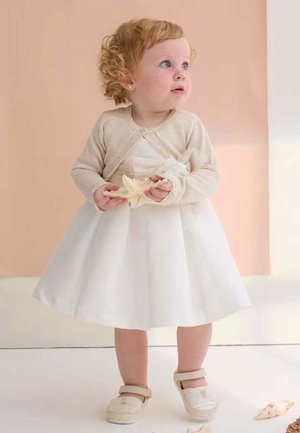 Bambina in abito bianco e cardigan beige che tiene una conchiglia, in piedi su sfondo color pesca chiaro, guarda di lato.