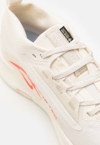 Chaussure de sport beige clair avec un dessus texturé, des accents orange prononcés, un col rembourré et des lacets plats. Marque sur la languette et sur le côté.