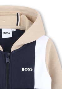 Veste à zip avec capuche en marine, crème et blanc. Fabriquée en coton doux, avec une texture côtelée et un détail de logo à l'avant.