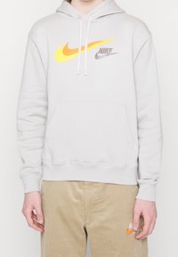 Ljusgrå hoodie med en känguruficka, prydd med djärva gula och orange Nike swoosh-logotyper och dragsnören. Bomullsmaterial, avslappnad passform.