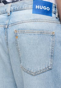 Jean en denim bleu clair avec des coutures visibles sur la poche arrière et une étiquette bleue portant la mention "HUGO" près de la ceinture.