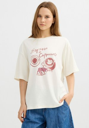 Joven mujer con camiseta blanca con ilustración de tazas de espresso, cappuccino y croissant, y vaqueros azules, de pie frente a un fondo liso.