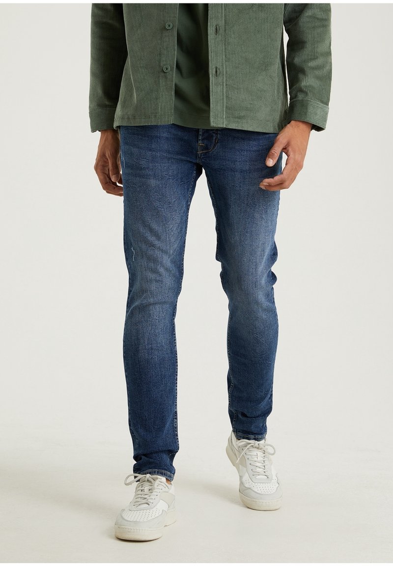 Jeans ajustados de mezclilla en azul oscuro, combinados con una chaqueta de pana verde y zapatillas blancas, con una superficie texturizada y cierre de botón.