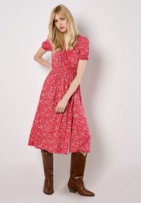 Robe midi rouge à fleurs avec des manches courtes bouffantes, une taille froncée et une jupe fluide. Portée avec des bottes de cowboy marron.