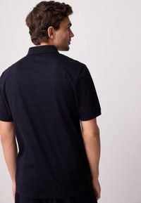 Temno modra polo majica s kratkimi rokavi, teksturiranim karo vzorcem in ovratnikom. Zadnji pogled prikazuje gladko tkanino in detajle toniranih šivov.