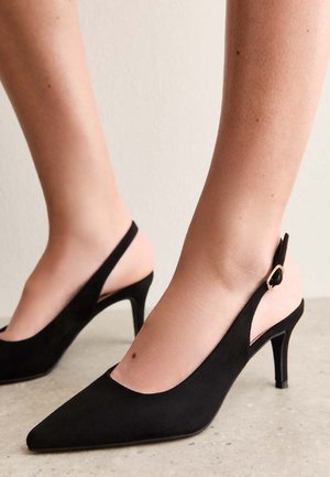 Czarne zamszowe sandały na obcasie typu slingback z zaostrzonymi noskami i średniej wysokości obcasami szpilkowymi, noszone na boso na neutralnym tle.