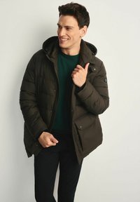 Ciemnozielona kurtka puchowa z kapturem, z zamkiem błyskawicznym, kieszeniami na zatrzaski oraz pikowanym wzorem. Nosi na sobie zielony sweter i czarne spodnie.