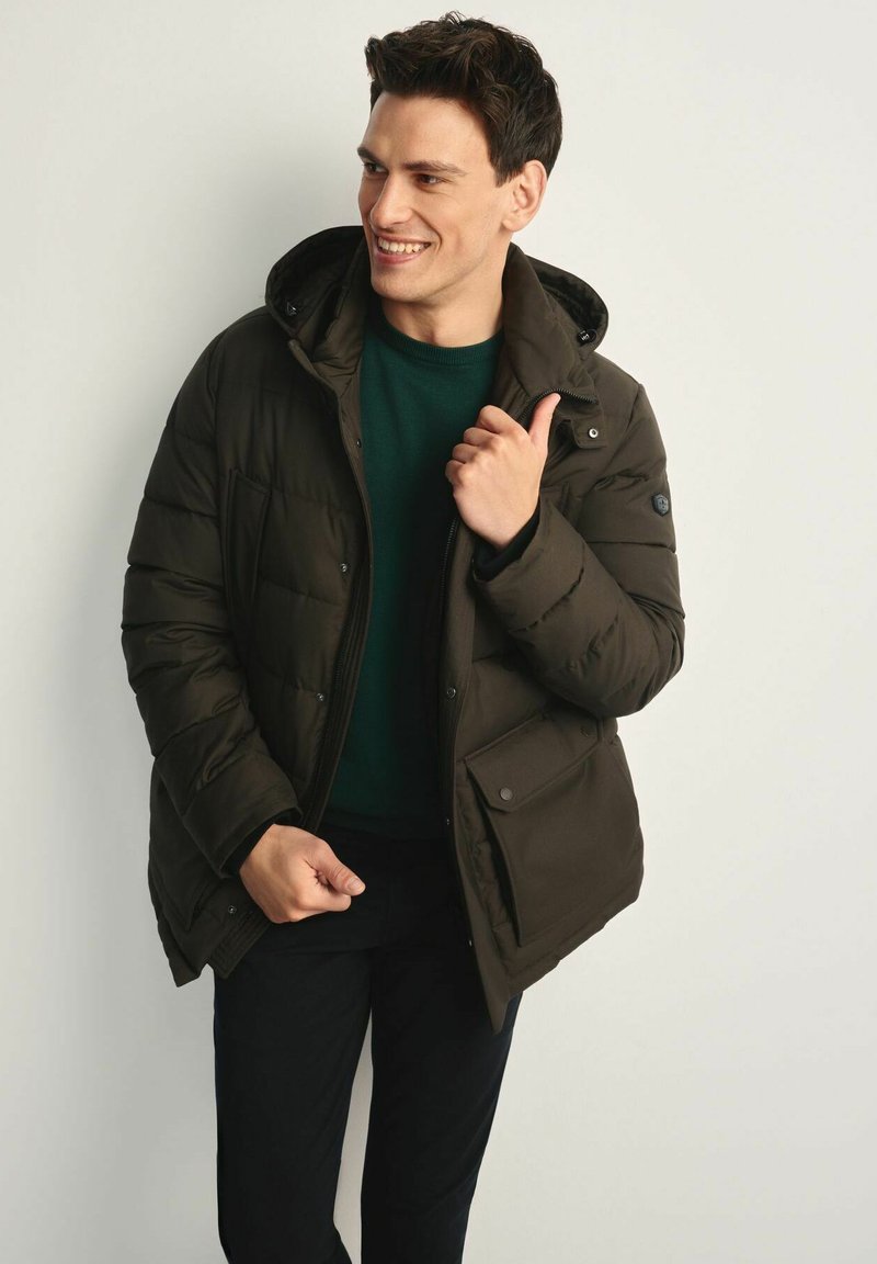 Ciemnozielona kurtka puchowa z kapturem, z zamkiem błyskawicznym, kieszeniami na zatrzaski oraz pikowanym wzorem. Nosi na sobie zielony sweter i czarne spodnie.