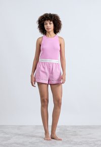 Ροζ ριγέ μπλουζάκι με αμάνικα συνδυασμένο με ανοιχτό ροζ κοντές παντελόνες με λευκή ζώνη. Το μοντέλο στέκεται ξυπόλυτο σε ανοιχτό χαλί, σε ουδέτερο φόντο.