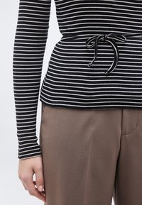 Μακρυμάνικη μπλούζα με ασπρόμαυρες ρίγες και δεσίματα στη μέση, συνδυασμένη με καφέ παντελόνια υψηλής μέσης σε άτομο που στέκεται.