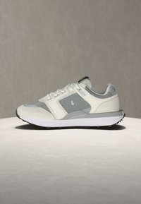 Zapatilla de deporte gris claro y blanca con paneles de malla, cordones blancos y un pequeño logotipo en el lateral, colocada sobre una superficie plana y neutra.