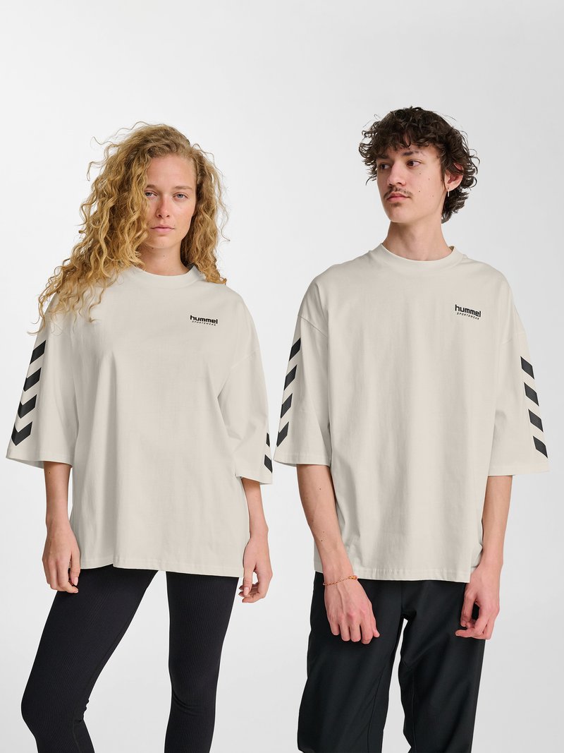 Deux jeunes adultes portant des t-shirts Hummel beige oversize avec des bandes chevron noires sur les manches, debout devant un fond blanc uni.