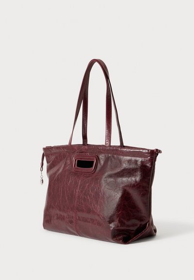 Sac cabas en cuir rouge foncé avec doubles bretelles et petite découpe rectangulaire pour la poignée sur un fond clair uni.