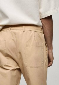 Bež hlače iz lahkotnega materiala z elastičnim pasom, dvema zadajnima žepoma in gladko teksturo. Tkanina se zdi zračna in sproščena.