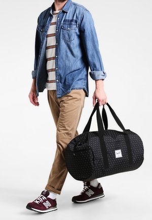 Man wearing denim shirt, striped t-shirt, tan pants, and maroon sneakers carrying a black duffel bag with patterned design by its handles.