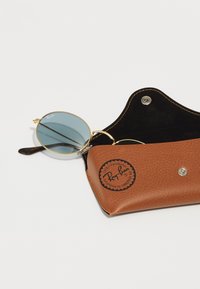 Gafas de sol de montura dorada en forma redonda con lentes azules, colocadas dentro de una funda de cuero texturizado marrón con un adhesivo de logotipo negro.