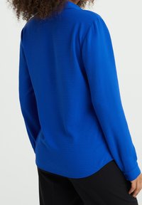 Långärmad skjorta i livlig blå färg, med en slät yta, krage och en rundad nederkant, i kombination med svarta byxor.