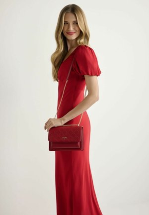 Kobieta w czerwonej sukience z bufiastymi rękawami, niosąca czerwoną pikowaną torebkę z złotym łańcuszkiem, uśmiechająca się i patrząca na bok.