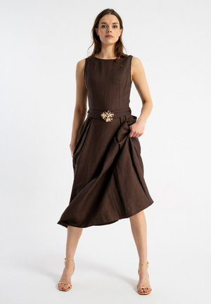 Kobieta w ciemnobrązowej sukience midi bez rękawów z ozdobnym paskiem, trzymająca spódnicę i nosząca złote sandały na wysokim obcasie.