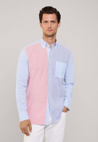 Camisa abotonada con rayas verticales; lado izquierdo rojo y blanco, lado derecho azul claro y blanco. Tiene un bolsillo en el pecho y un cuello.