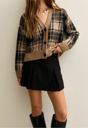 Femme portant un cardigan marron à carreaux boutonné, une mini-jupe plissée noire, des colliers superposés et des bottes noires montant jusqu'aux genoux.