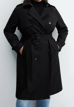 Schwarzer doppelt geknöpfter Trenchcoat mit Gürtel, mit geknöpften Ärmelsäumen, getragen über schwarzem Top und blauer Jeans, mit Händen in den Manteltaschen.