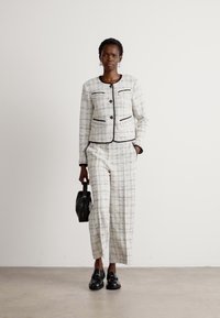 Costume en tweed blanc et gris avec bordure noire, comprenant une veste ajustée et un pantalon à jambes larges. Le mannequin tient un petit sac à main noir et porte des chaussures noires.