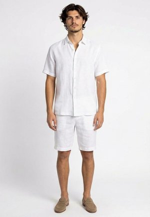 Uomo in piedi che indossa una camicia bianca a maniche corte con bottoni, pantaloncini bianchi al ginocchio e mocassini beige su uno sfondo semplice.