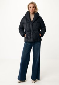 Doudoune noire avec un col haut, un design matelassé et des manches longues. Associée à un jean en denim bleu à jambes larges et des chaussures beiges.