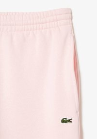 Pantalones de chándal rosas con cinturilla elástica, que presentan un pequeño logo de cocodrilo verde en la pierna izquierda y un acabado suave y texturizado.