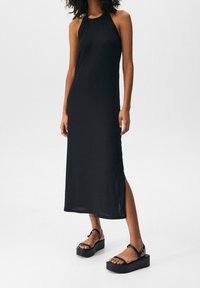 Femme portant une robe midi noire sans manches à côtes avec une fente latérale et des sandales compensées noires, debout sur un fond blanc.