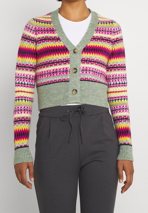 Femme portant un cardigan court avec des motifs géométriques multicolores et une bordure verte, associé à un pantalon noir avec cordon de serrage et un haut blanc.