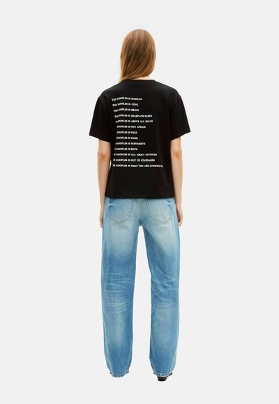 Persoon met lang haar die een zwart T-shirt met witte tekst op de rug draagt en losse lichtblauwe jeans, staand tegen een witte achtergrond.