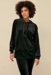 Zwarte velours hoodie met een voorzak, lange mouwen en een verstelbare trekkoord. Zachte textuur en een relaxte pasvorm. Gepaard met bijpassende broek.