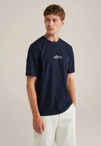 Navyblaue Baumwoll-T-Shirt mit Rundhalsausschnitt, kurzen Ärmeln und weißem Drucktext auf der Brust. Das Model trägt hellfarbene Hosen.