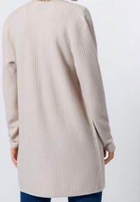 Haut à manches longues beige avec texture côtelée et ourlet arrondi. Matière douce, coupe décontractée, et fentes latérales pour le mouvement et le confort.