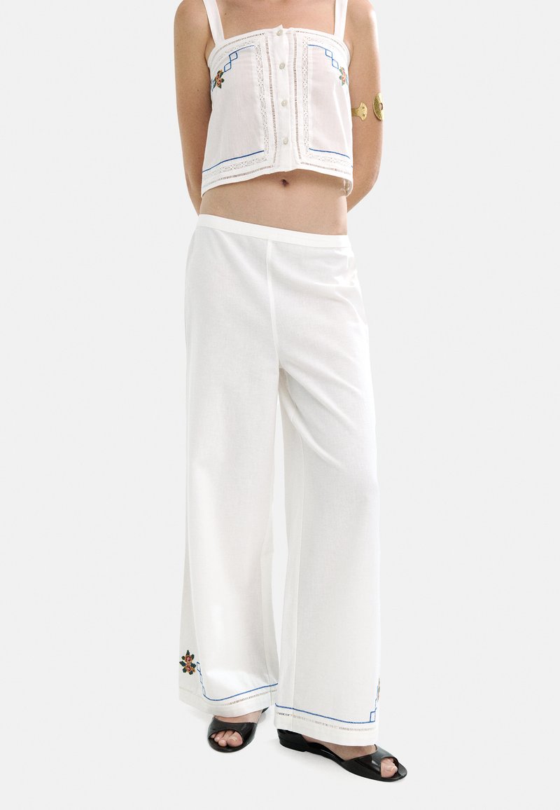 Biały top krótki z kwiatowym haftem, guzikami i niebieskimi akcentami, zestawiony z luźnymi białymi spodniami z kwiatowym wzorem na dolnej części.