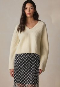 Krémový rebrovaný pletený sveter s výstrihom do V a skráteným strihom, skombinovaný s čiernou sukňou s bielymi kvetinovými vzormi a priesvitným čipkovým lemom.