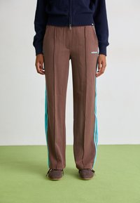 Braune Nadelstreifen-Hosen mit türkisfarbenen Seitenstreifen, im geraden Schnitt. Getragen mit dunklem Schuhwerk und einem marineblauen Pullover.