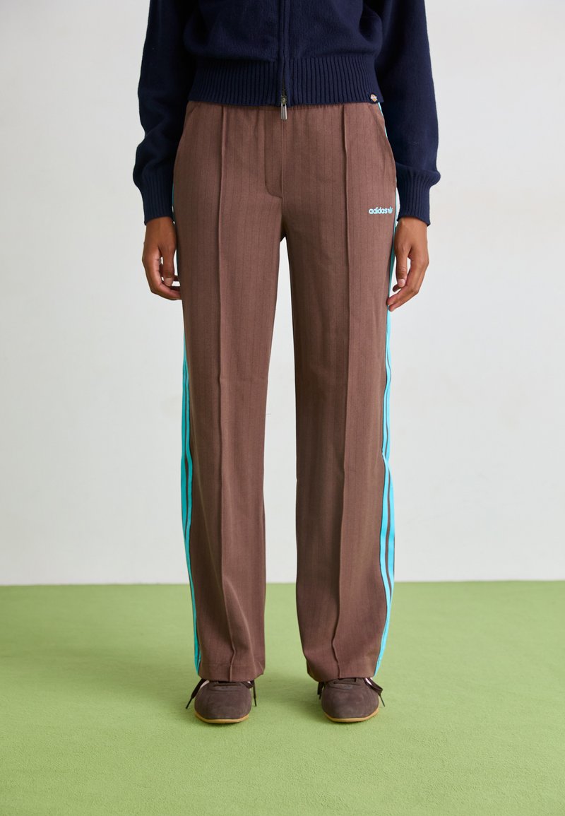 Braune Nadelstreifen-Hosen mit türkisfarbenen Seitenstreifen, im geraden Schnitt. Getragen mit dunklem Schuhwerk und einem marineblauen Pullover.