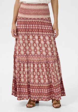 Woman wearing a long, patterned skirt with red and beige geometric and floral designs, paired with tan open-toe sandals and a white top.