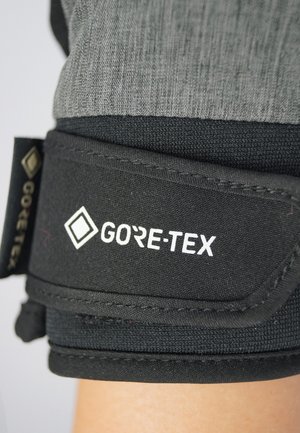 Black GORE-TEX fabric with a smooth texture, featuring a gray textile upper and adjustable strap, showcasing a prominent logo in white.
