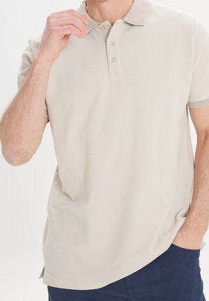 Uomo che indossa una polo beige a maniche corte con tre bottoni, una mano vicino al colletto, l'altra nella tasca, pantaloni blu navy visibili.