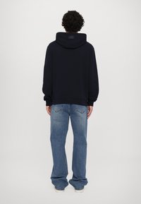 Felpa blu navy con tasca a marsupio e logo sulla schiena, abbinata a jeans larghi di un blu chiaro con tasche posteriori e cuciture visibili.