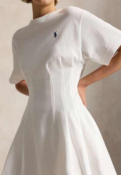 Γυναίκα με λευκό φόρεμα κοντομάνικο με εφαρμοστή μέση και μπλε κεντημένο λογότυπο πόλο στο στήθος, στέκεται με τα χέρια στη μέση.