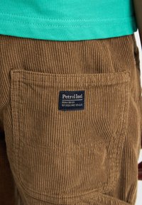Pantaloni in corduroy marrone con una tasca posteriore dotata di un'etichetta blu che dice "Petrol Ind." Il tessuto presenta righe e texture visibili.