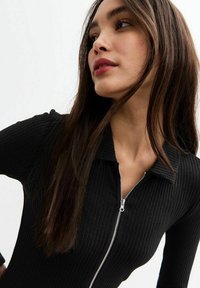 Svart rippad bodysuit med dragkedja, med kragkrage och långa ärmar; slät textur och figurnära design.