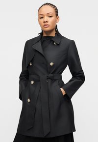 Γυναίκα που φοράει μαύρο trench coat με ζώνη και μεγάλα κουμπιά, ποζάροντας με τα χέρια στις τσέπες μπροστά σε λευκό φόντο.