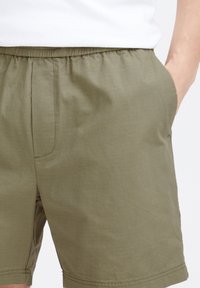 Õliverohelised lühikesed püksid, valmistatud kerge kangaga, mis on mugava lõikega, elastse taljega ja küljetaskutega. Õrn tekstuur on nähtav.